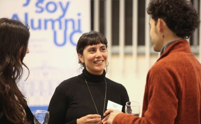 Fotografía de tres personas conversando, se ve la cara de una de ella sonriendo, y al fondo difuminado se ve en grande #YoSoyAlumniUC