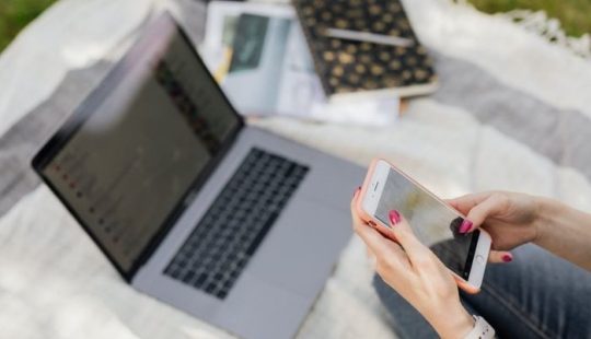 Fotografía de las manos de una persona sosteniendo un celular sobre una manta sobre pasto, encima un computador portátil, una libreta, un lápiz y otros elementos similares