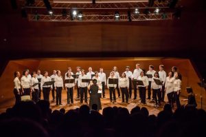 Fotografía del Coro Alumni UC hace años, cantando en un escenario vestidos con camisa blanca y pantalón negro