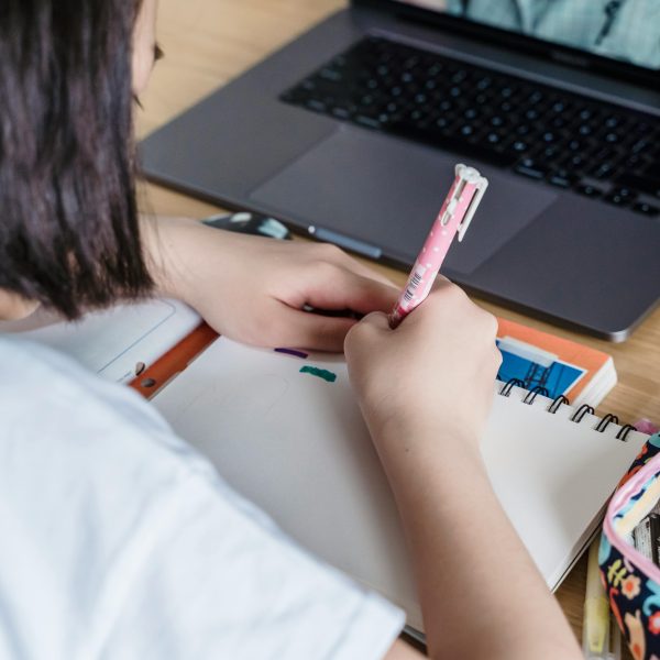 Fotografía de una niña escribiendo en un cuaderno frente a un computador portátil