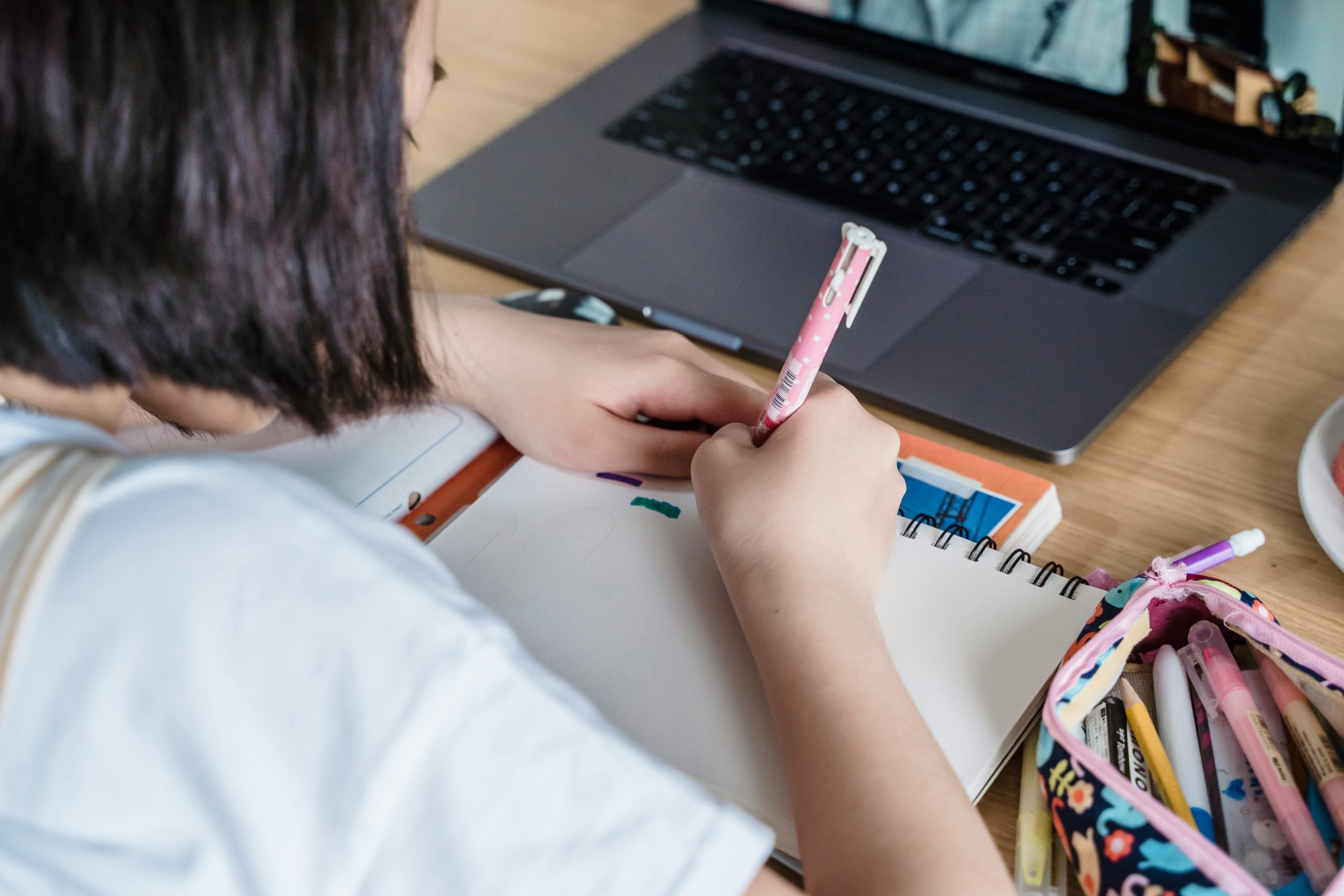 Fotografía de una niña escribiendo en un cuaderno frente a un computador portátil