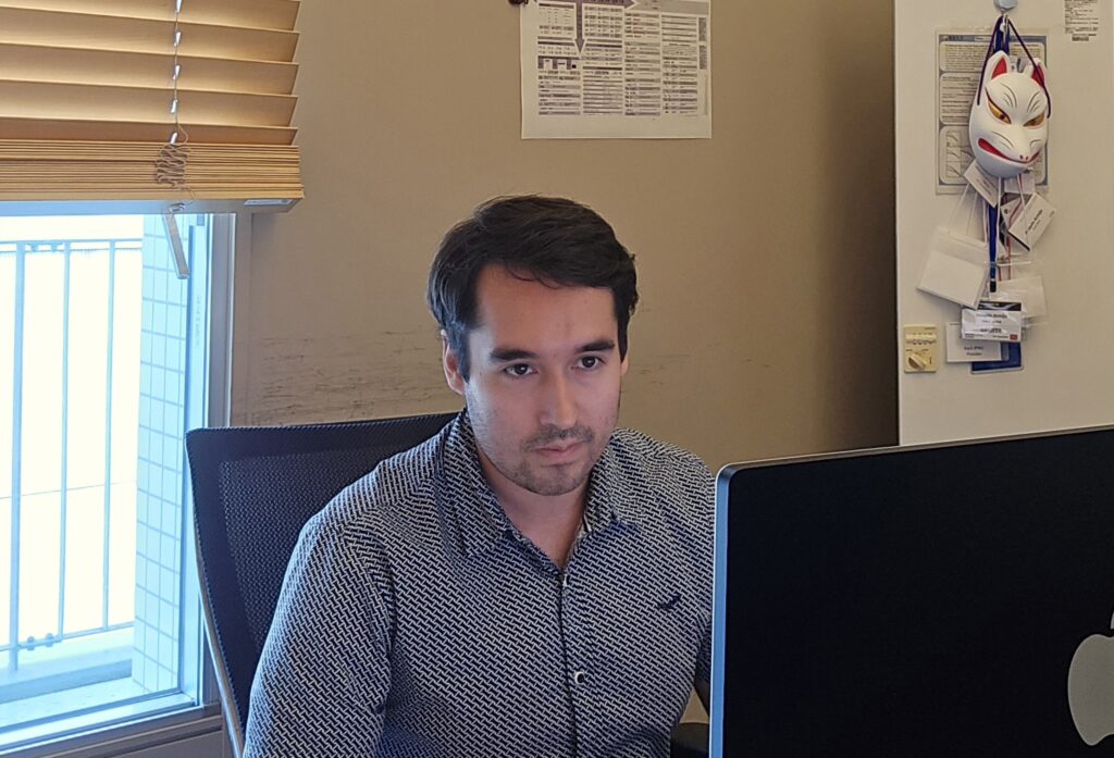 Fotografía de Joaquín Armijo frente a un computador en el entorno de una oficina individual, de fondo se ve una máscara japonesa y una ventana