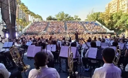 Fotografía de una orquesta vista desde atrás y de fondo cientos de personas en el público