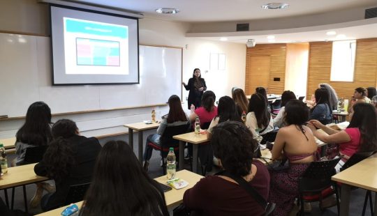 Fotografía de una sala de clases, se ven decenas de personas sentadas y una persona en frente proyectando una presentación