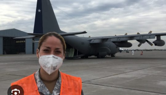 Fotografía de María en una base aérea, frente a un avión estacionado en la pista.