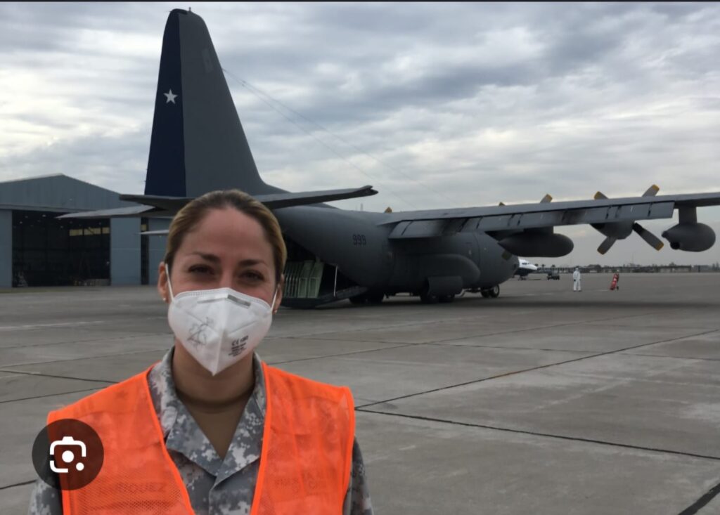 Fotografía de María de los Ángeles Henríquez con mascarilla, traje militar, un chaleco reflectante naranjo y de fondo un gran avión modelo Hércules