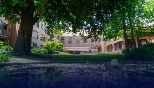 Fotografía de una sede de Senior suits, se ve de fondo un gran edificio y frente a éste mucho verde y una pequeña laguna