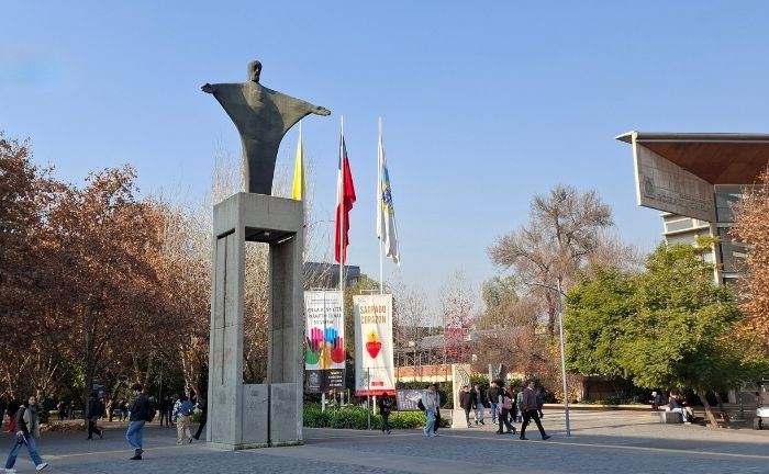 Fotografía del frontis del Campus San Joaquín se ve el gran Cristo junto a las banderas de Chile y de la Universidad Católica, y gente caminando en distintas direcciones.