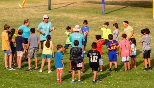 Fotografía de un grupo de decenas de niños y niñas junto a monitores en una cancha de pasto