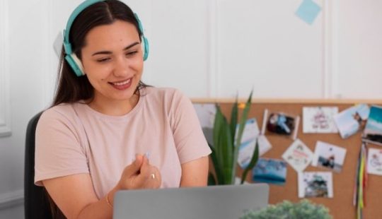 Fotografía de una mujer sonriendo con unos audífonos grandes tipo cintillo color celeste, mirando un computador, de fondo se ven unas fotografías difuminadas en la pared, y una planta