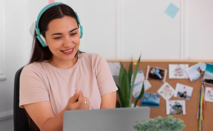 Fotografía de una mujer sonriendo con unos audífonos grandes tipo cintillo color celeste, mirando un computador, de fondo se ven unas fotografías difuminadas en la pared, y una planta