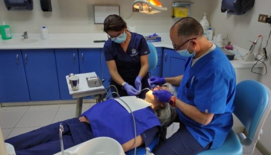 Fotografía de una de las consultas de la Clínica Dental de la Dirección de Personas UC, se ve una paciente acostada mostrando los dientes, un dentista y una asistente, ambos con mascarilla e implementos de odontología