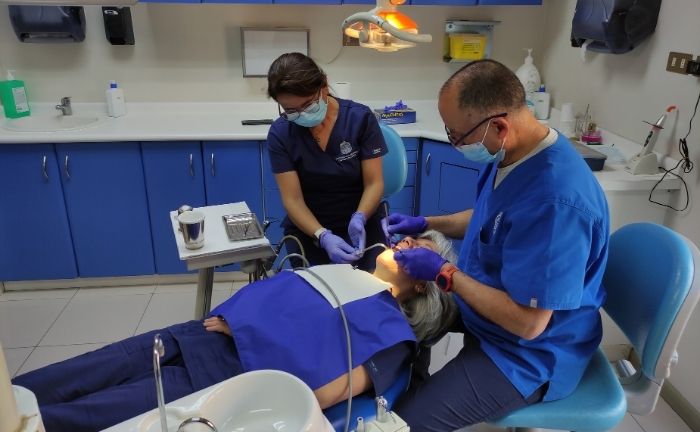 Fotografía de una de las consultas de la Clínica Dental de la Dirección de Personas UC, se ve una paciente acostada mostrando los dientes, un dentista y una asistente, ambos con mascarilla e implementos de odontología