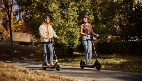 Fotografía de dos personas en un scooter cada una, andando en un camino de cemento alrededor de una zona de pasto y árboles
