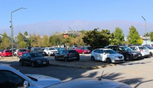Fotografía de uno de los estacionamientos del Campus San Joaquín UC, se ven decenas de autos estacionados y varios focos de luz, de fondo se ve la cordillera