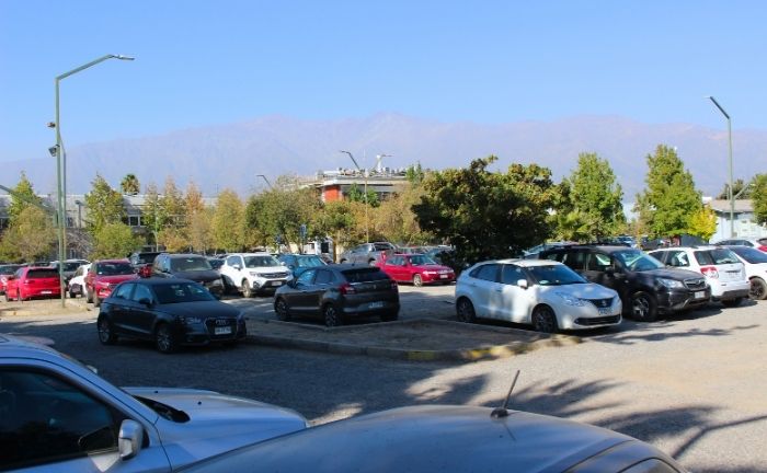 Fotografía de uno de los estacionamientos del Campus San Joaquín UC, se ven decenas de autos estacionados y varios focos de luz, de fondo se ve la cordillera