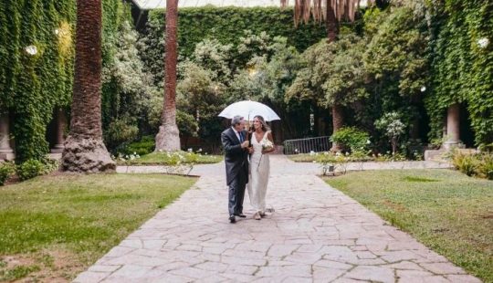 Fotografía de una novia junto a su padre en uno de los patios del Campus Oriente UC, se ve un camino de piedras, pasto y troncos de palmeras chilenas