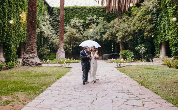 Fotografía de una novia junto a su padre en uno de los patios del Campus Oriente UC, se ve un camino de piedras, pasto y troncos de palmeras chilenas