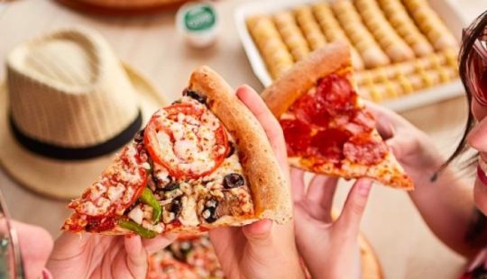 Fotografía de las manos de dos personas sosteniendo dos trozos de pizza, en el fondo se ve una mesa con palitos de ajo, salsa de ajo de Papa John's y un sombrero