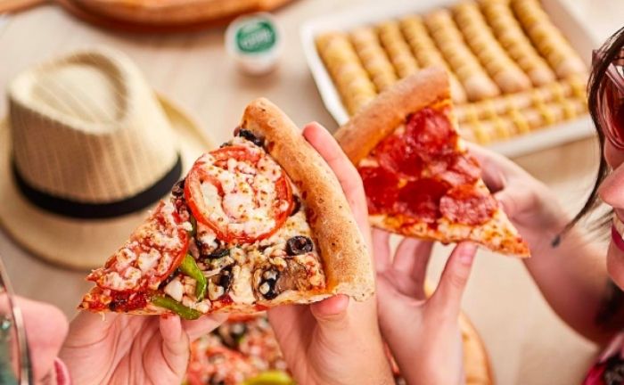 Fotografía de las manos de dos personas sosteniendo dos trozos de pizza, en el fondo se ve una mesa con palitos de ajo, salsa de ajo de Papa John's y un sombrero