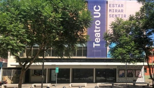 Fotografía del frontis del Teatro UC, se ve un gran telón con el nombre del teatro, y a un costado escrito "Estar, mirar, sentir"