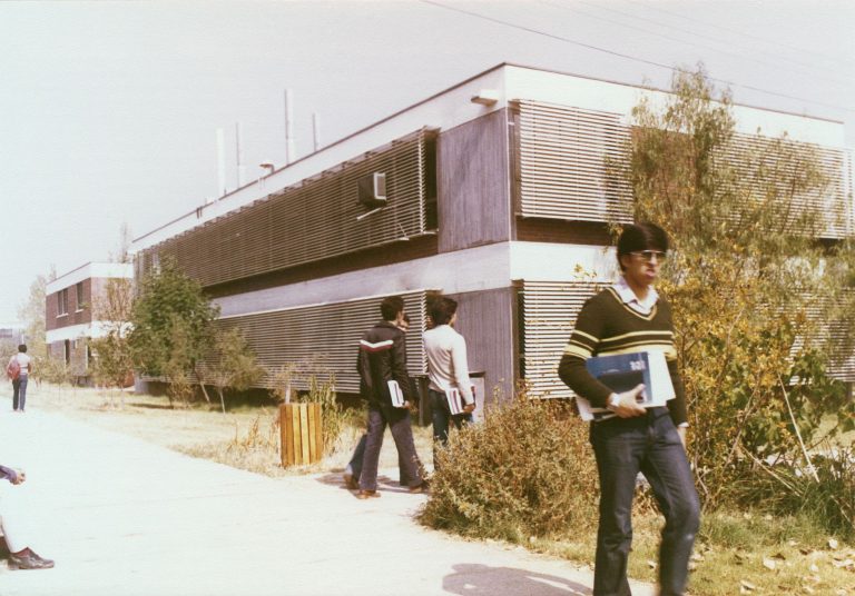 Fotografía de estudiantes llevando libros en las manos, caminando por un campus universitario frente a un edificio académico de dos pisos, con árboles y áreas verdes alrededor.
