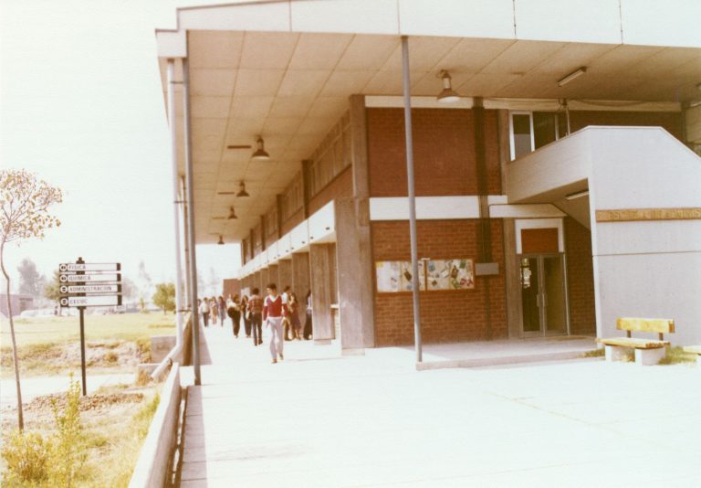Personas caminando por un campus universitario, frente a un edificio académico de dos niveles, con pasillos exteriores y áreas abiertas.