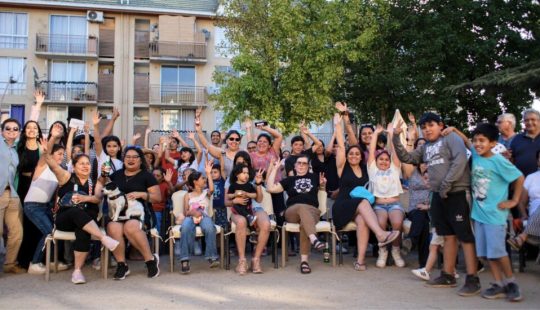 Fotografía de un grupo numeroso de personas adultas y niños reunidos al aire libre, sentados y de pie,frente a edificios residenciales.