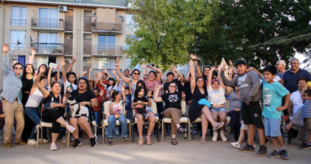 Fotografía de un grupo numeroso de personas adultas y niños reunidos al aire libre, sentados y de pie,frente a edificios residenciales.