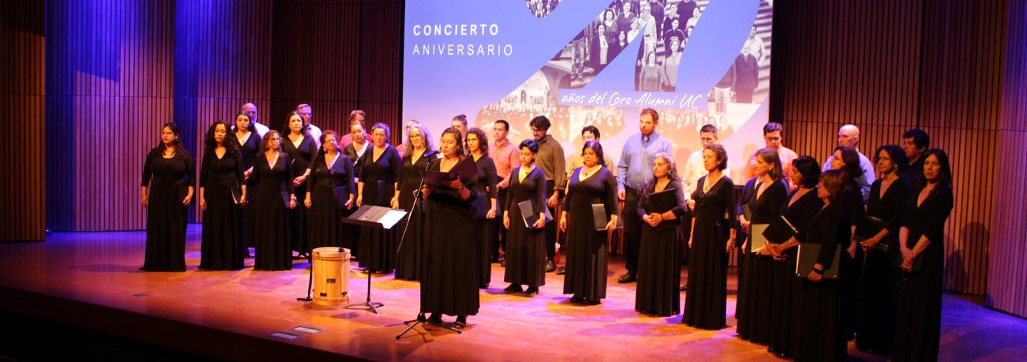 Fotografía de un grupo coral vestidos de negro en un auditorio, de fondo se ve una gráfica proyectada que dice 20 años Coro Alumni UC, y el número 20 con un collage de fotos
