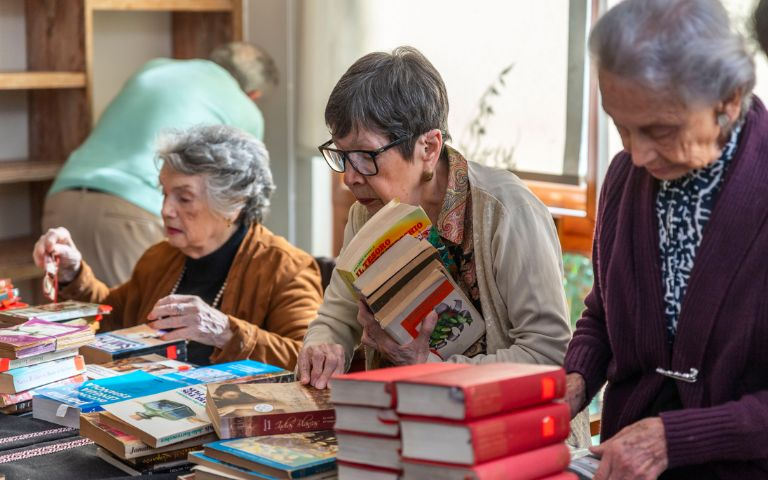 Tres mujeres adultas mayores revisando y organizando pilas de libros sobre una mesa en un espacio interior.