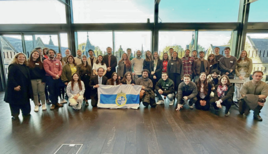Fotografía de un grupo de más de 30 personas sosteniendo la bandera de la Universidad Católica