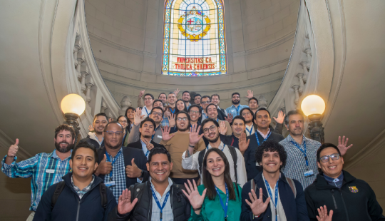 Fotografía de un grupo de decenas de personas sonriendo y alcanzo la palma de la mano