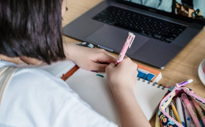 Fotografía de una niña escribiendo en un cuaderno frente a un computador portátil